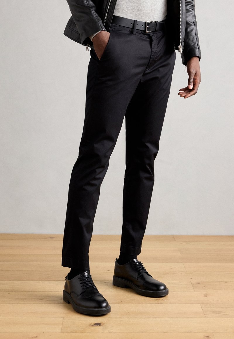 Black tailored trousers with a smooth texture, slim fit, and a matching black leather belt. Worn with black lace-up shoes.