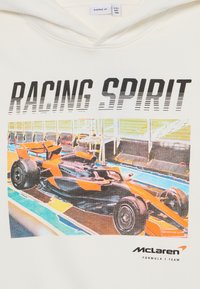 Kreem värvi kapuutsiga, millel on värvikas graafika McLaren Formula 1 autost ja tekst "RACING SPIRIT" üleval, detailsete aktsentidega oranžis ja mustas.