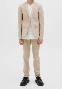 Traje de lino beige claro con chaqueta y pantalones a juego, combinado con una camisa blanca abotonada. Zapatos negros completan el conjunto.