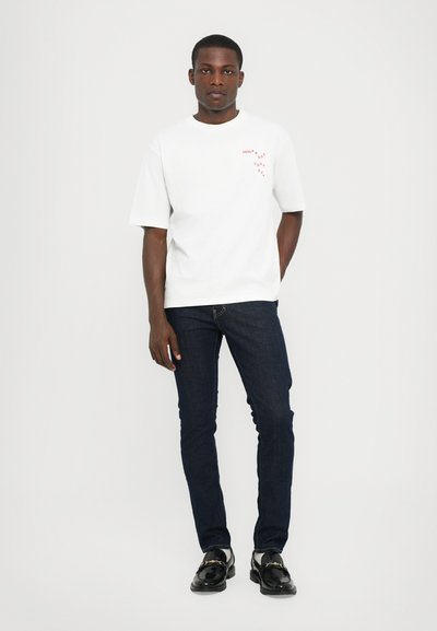 White cotton t-shirt with a round neckline, short sleeves, and red circular graphic print on the chest. Paired with dark blue slim-fit jeans.