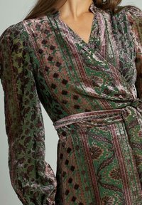 Grünes Wickelkleid mit langen Puffärmeln, das florale und geometrische Muster in Grüntönen, Gold und Burgunderrot aufweist.