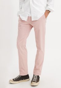 Uomo con pantaloni rosa chiaro, scarpe da ginnastica nere alte e una maglietta bianca a maniche lunghe, con le mani nelle tasche, in piedi su uno sfondo bianco.
