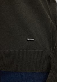 Maglione lavorato a maglia verde scuro con orlo a costine; presenta un'etichetta con logo metallico che riporta "JOOP!" attaccata al tessuto.