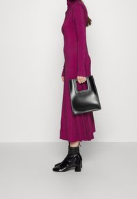 Fuchsia long-sleeve dress with textured pattern, paired with a black patent leather handbag featuring a handle and squared shape, and ankle boots.