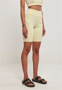 Pantaloni sportivi attivi di colore giallo chiaro con vita alta, caratterizzati da un design aderente e pannelli laterali a contrasto. Abbinati a sandali neri con slide.