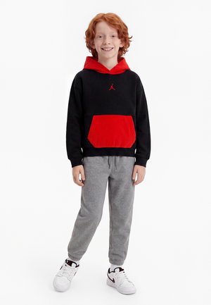 Garçon roux souriant portant un sweat à capuche noir et rouge, un pantalon de survêtement gris et des baskets blanches sur un fond blanc uni.