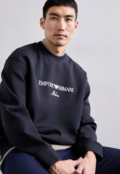 Sweatshirt bleu marine avec un col rond, arborant le texte blanc "EMPORIO ARMANI Milano". Tissu doux avec une coupe décontractée et des manches longues.