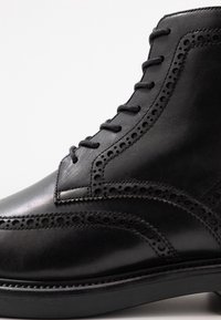 Černé kožené kotníkové boty s ozdobným prošíváním, kulatou špičkou a šněrovacím designem. Podešev je texturovaná pro lepší přilnavost a stabilitu.