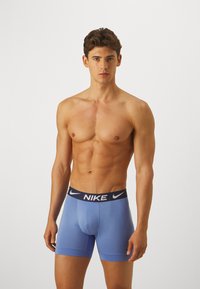 Plave Nike bokserske gaće s tamnoplavim elastičnim pojasom na kojem se nalazi logo. Tkanina izgleda glatko, usko i rastezljivo, pružajući udoban dizajn.