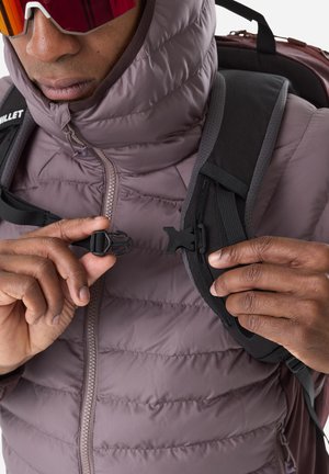 Personne portant une doudoune mauve et des lunettes de soleil, attachant la boucle de la sangle noire de son sac à dos sur la poitrine.