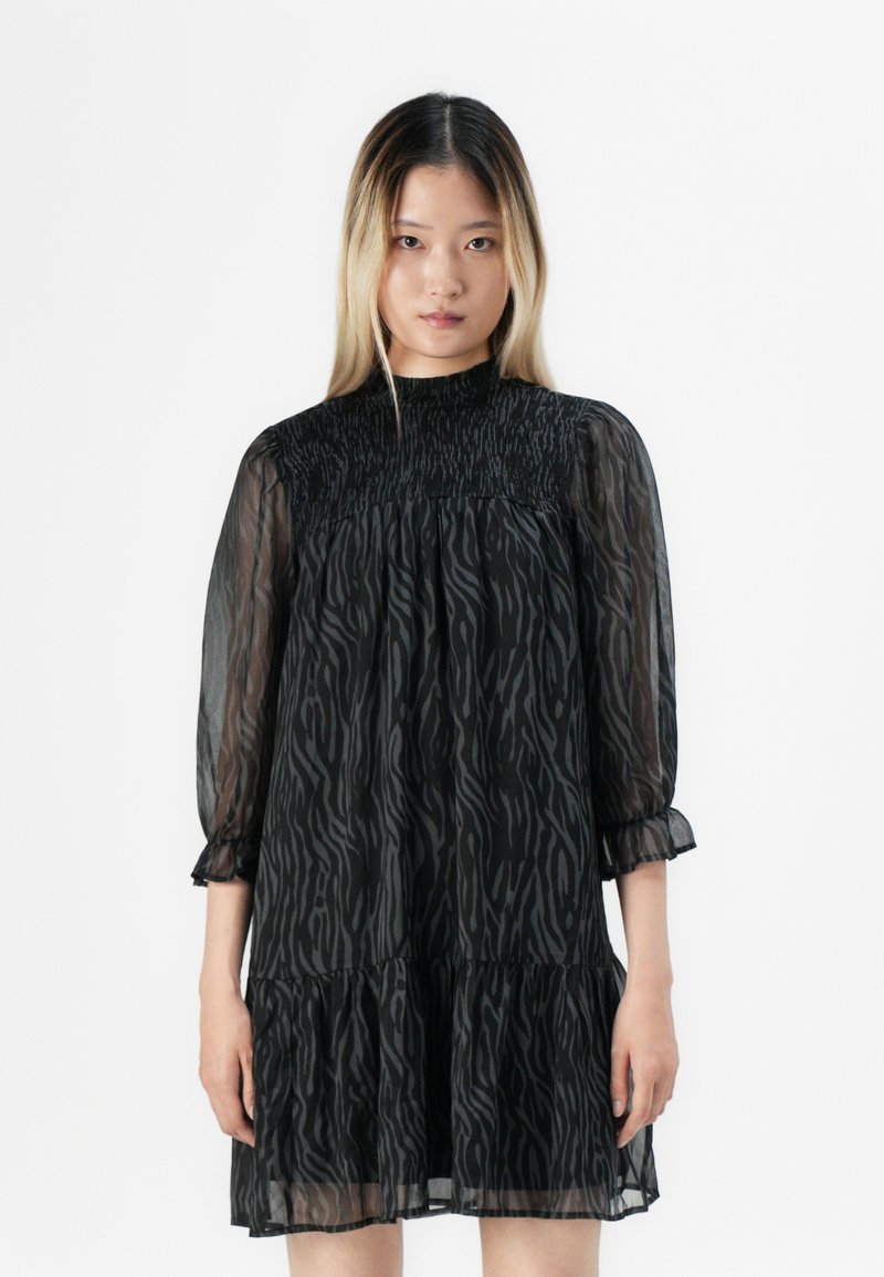 Jovem mulher com cabelo comprido e liso veste um vestido preto, transparente, com padrão, decote alto e mangas três quartos, contra um fundo branco.