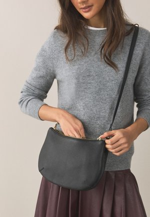 Femme en pull gris et jupe bordeaux tenant et dézippant un sac bandoulière en cuir noir avec une fermeture éclair dorée.