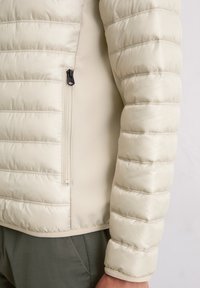 Beżowa pikowana kurtka puchowa z poziomym przeszyciem; wyposażona w kieszeń na zamek oraz elastyczne mankiety zapewniające dopasowanie. Miękki, lekki materiał.