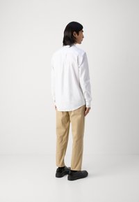 White long-sleeve shirt with a curved hem, button cuffs, and a pleat at the back, paired with beige trousers and black shoes.
