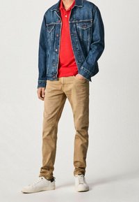 Chaqueta de mezclilla, polo rojo, pantalones beige y zapatillas blancas. La chaqueta tiene dos bolsillos en el pecho y detalles de botones. Tejido de mezcla de algodón y corte informal.