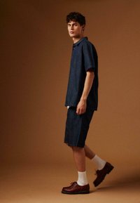 Jeune homme marchant, portant une chemise à manches courtes bleu foncé et un short assorti, des chaussettes blanches et des chaussures en cuir marron, sur un fond brun uni.