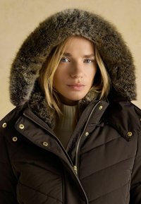 Hnedá péřová bunda s kapucňou s kožúškom, so zlatými detailmi, prešívaným dizajnom a vysokým golierom. Pod ňou je ľahký sveter.