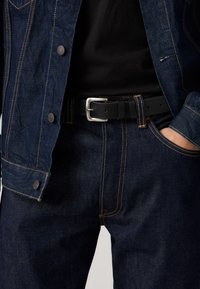 Veste et pantalon en jean bleu foncé avec coutures orange, équipés d'une ceinture en cuir noir avec une boucle argentée. Matériau texturé.