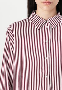 Gros plan d'une personne portant une chemise blanche à rayures verticales rouge foncé et boutons blancs, montrant le col et une partie du visage.