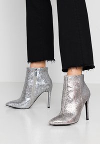 Botines de tobillo plateados con glitter, punta afilada y tacón stiletto alto. Cuentan con una cremallera lateral para el cierre y una textura suave y brillante.