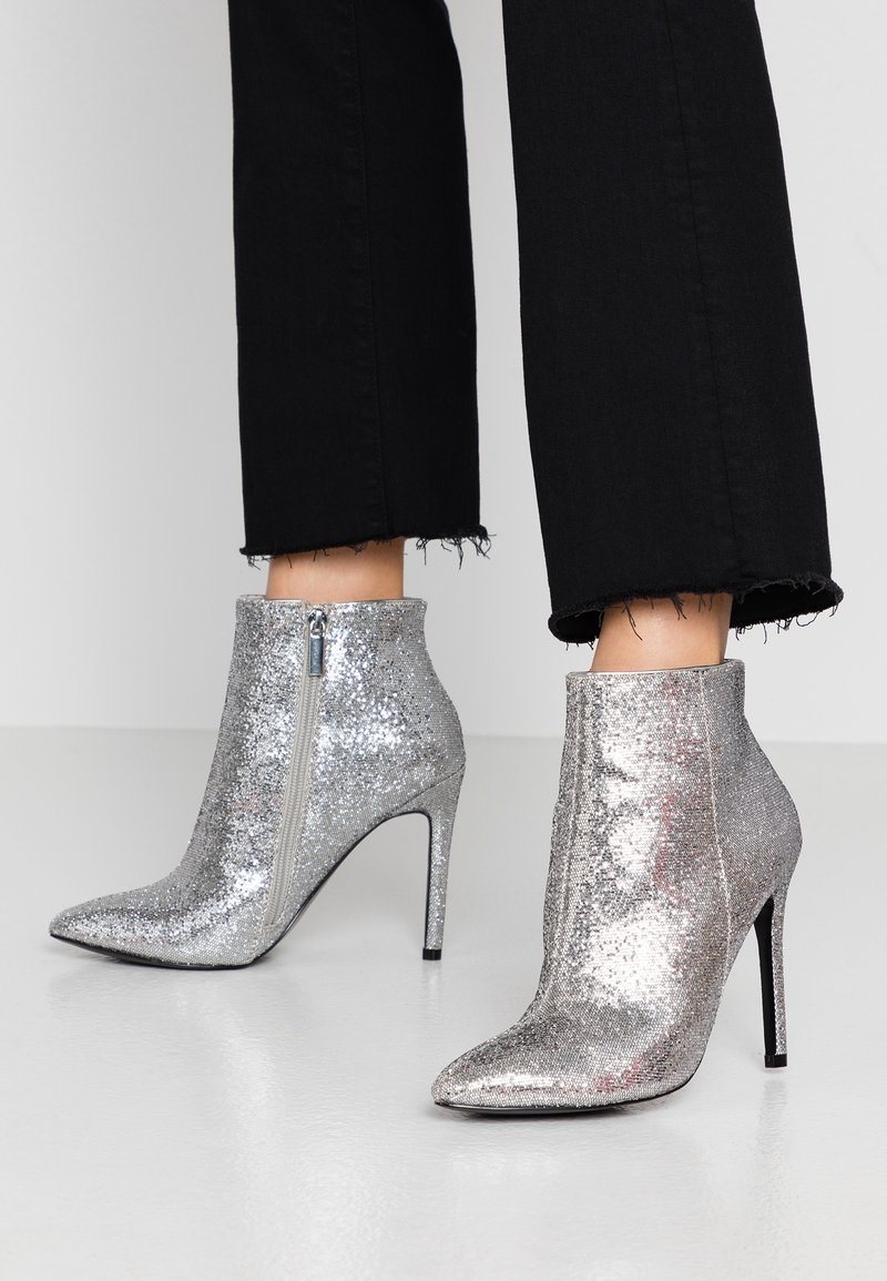 Botines de tobillo plateados con glitter, punta afilada y tacón stiletto alto. Cuentan con una cremallera lateral para el cierre y una textura suave y brillante.