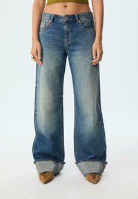 Džínsy s wide-leg strihom v stredne modrom umývaní, s jemným vyblednutím, vysokým pásom a rolovanými manžetami. Styled s špicatými hnedými topánkami.