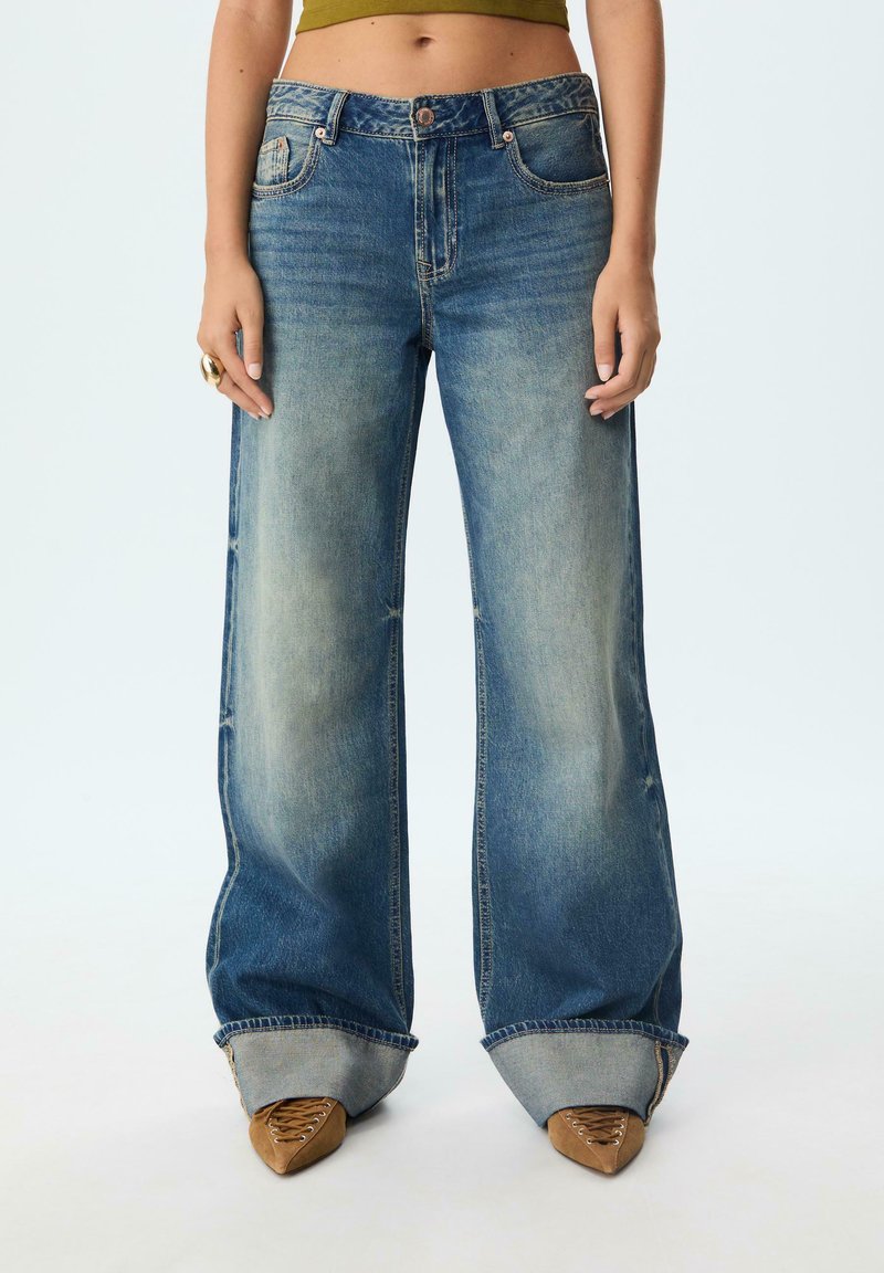 Džínsy s wide-leg strihom v stredne modrom umývaní, s jemným vyblednutím, vysokým pásom a rolovanými manžetami. Styled s špicatými hnedými topánkami.