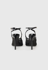 Sandales à talons hauts noires avec une finition laquée brillante, présentant des brides croisées au niveau des chevilles et un détail en maille sur les côtés.