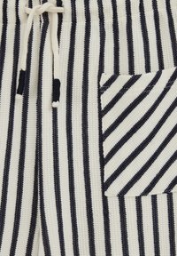 Tissu en maille blanche avec des rayures noires verticales, doté d'un cordon de serrage avec des embouts noirs et d'une poche à rayures diagonales.