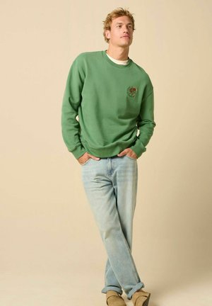 Junger Mann trägt grünes Sweatshirt mit Blumenstickerei, hellblaue Jeans und beige Schuhe, steht mit den Händen in den Taschen vor einfachem Hintergrund.