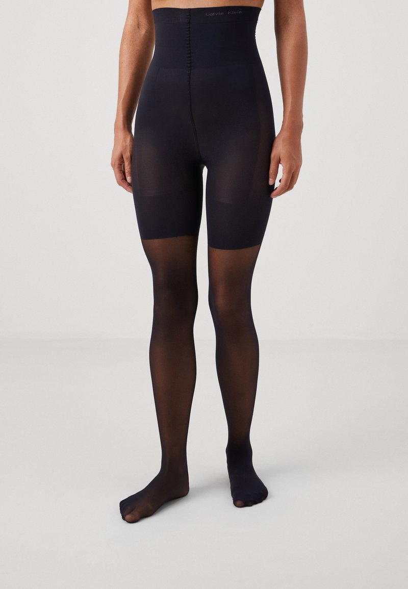 High-waisted black tights made of sheer material, featuring a smooth texture and opaque sections around the thighs. Subtle branding at the waistband.
