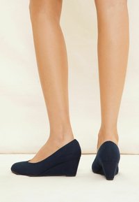 Tacchi a zeppa in suede blu navy con punta tondeggiante; presentano una texture liscia e un design a zeppa inclinata, offrendo una silhouette moderna.