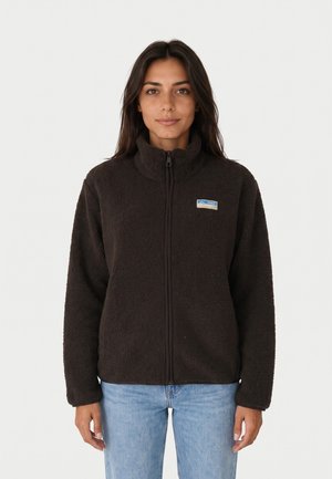 Mujer con chaqueta de forro polar negra con cremallera y un pequeño parche azul y blanco en el pecho, combinada con vaqueros azul claro.