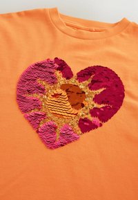 Tissu orange avec un motif en forme de cœur composé de sequins réversibles rose, rouge, or et orange disposés en motif de rayons de soleil.