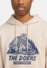 Beige hoodie featuring a blue graphic of a woodworker with trees. Drawstring details and soft fabric texture. Text reads "THE DOERS."