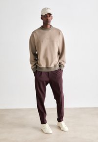 Man wearing beige cap, taupe sweatshirt, maroon pleated pants, and white sneakers standing against plain light background.