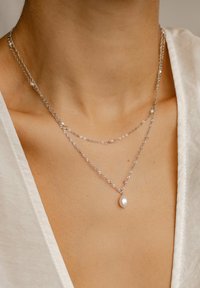 Srebrne naszyjniki warstwowe z skręconymi łańcuchami, z białą perłową zawieszką i drobnymi perłowymi akcentami. Teksturowane materiały i delikatny design.