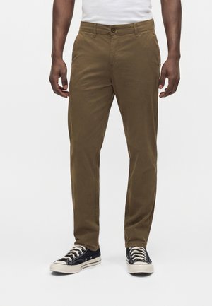 Homme portant un pantalon marron slim et des baskets basses noires, debout devant un fond blanc uni.