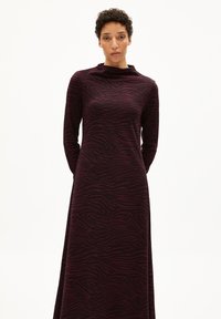 Frau in einem langärmeligen, hochgeschlossenen, dunkelvioletten Kleid mit schwarzen Zebrastrifen, die vor einem schlichten weißen Hintergrund steht.