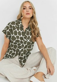 Camicia verde con un motivo geometrico bianco, maniche corte, chiusura a bottoni frontale e una singola tasca; abbinata a pantaloni di colore chiaro.