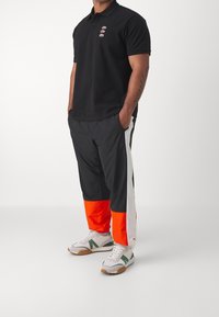 Muž v černém polo tričku se třemi malými logy, v černých sportovních kalhotách s bílými a oranžovými panely, v bílých teniskách se zelenými detaily, stojící s rukama v kapsách.