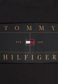 Camiseta negra con un panel de logo horizontal que lleva "TOMMY HILFIGER" en dorado y un emblema de color. Hecha de algodón con una textura suave.