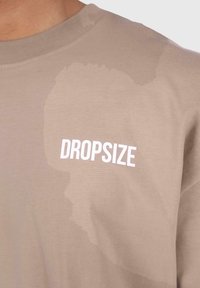Bež pamučna majica s bijelim grafikom "DROPSIZE" na prednjoj strani i suptilnim uzorkom u svjetlijoj nijansi bež boje.