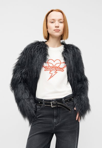 Μαύρο μπουφάν από ψεύτικη γούνα με μακριά μανίκια, φορεμένο πάνω από λευκό μπλουζάκι με κόκκινο γραφικό σχέδιο, σε συνδυασμό με σκούρο τζιν και ζώνη με τρουκς.