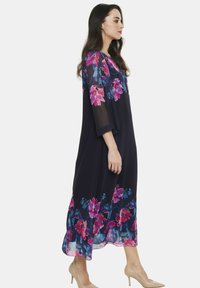 Granatowa sukienka maxi w kwiaty z przezroczystymi rękawami, z różowymi i niebieskimi wzorami kwiatowymi u dołu. Lekki materiał z luźnym krojem.
