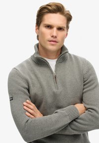 Grauer Zip-up-Pullover mit geripptem Kragen und Bündchen. Verfügt über einen halben Reißverschluss auf der Vorderseite, ein strukturiertes Strickmuster und ein Etikett am linken Ärmel.