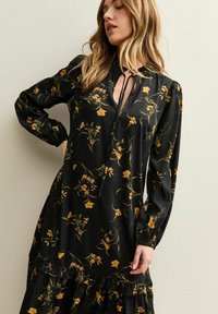 Zwarte bloemenjurk met gele bloemen, lange mouwen, V-hals met strikdetail en gelaagde rucheszoom. Gemaakt van gladde stof met een zachte textuur.