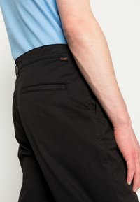 Schwarze Hosen mit einer glatten Textur, ausgestattet mit einer Gesäßtasche und einem kleinen orangefarbenen Etikett. Kombiniert mit einem hellblauen Hemd.