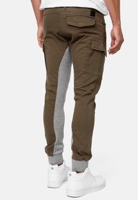 Cargo pants featuring brown cotton material with grey jersey inset, elastic cuffs, side pockets, and a back patch pocket. White sneakers shown.