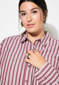 Bordeaux en witte gestreepte shirt met knoopsluiting, zichtbare borstzak en ronde knopen; model met ringen poseert met handen tegen de stof.